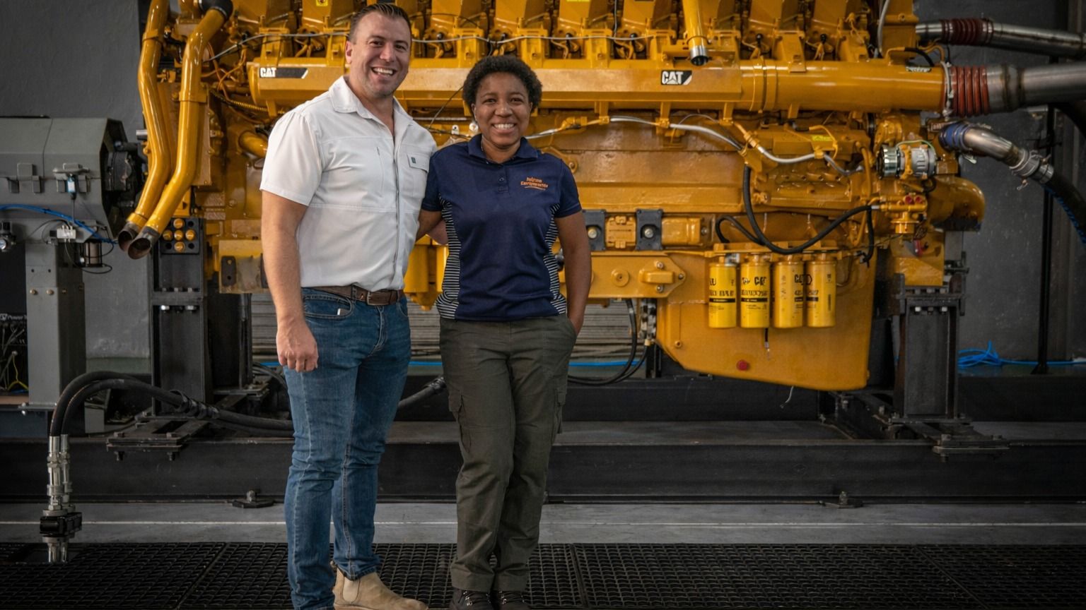 A smiling man and woman stand in front of a large yellow Caterpillar industrial engine.
